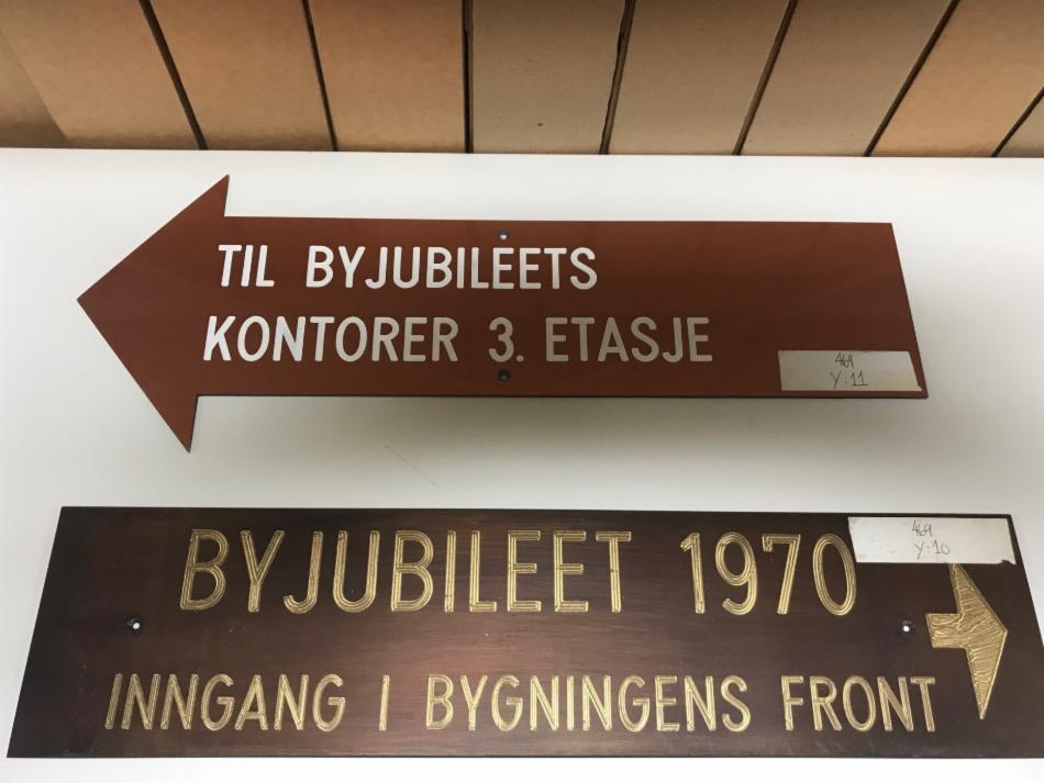 To av mange gjenstander i arkivet etter byjubileumskomiteen. Et skilt i plast til innendørs bruk, og et i bronse til å vise veien på utsiden av rådhuset. Foto: Kjerstin Kragseth.